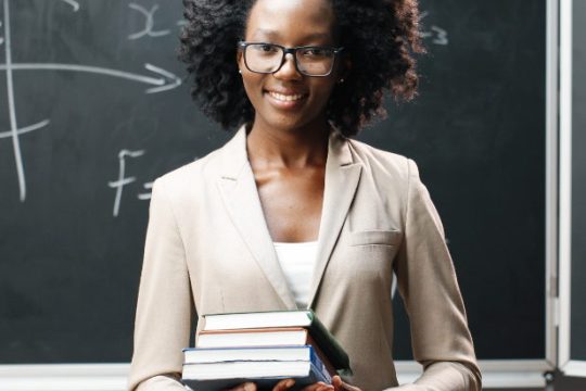 portrait-young-african-american-female-teacher-glasses-looking-camera-classrom-holding-textbooks-blackboard-with-formulas-background-schooling-concept-books-hands-woman (1)