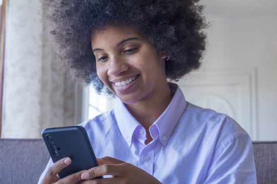 Smiling millennial african american woman using mobile phone at home. Happy black lady with curly hairstyle text messaging or chatting on smartphone. Young female browsing social media content on cellphone